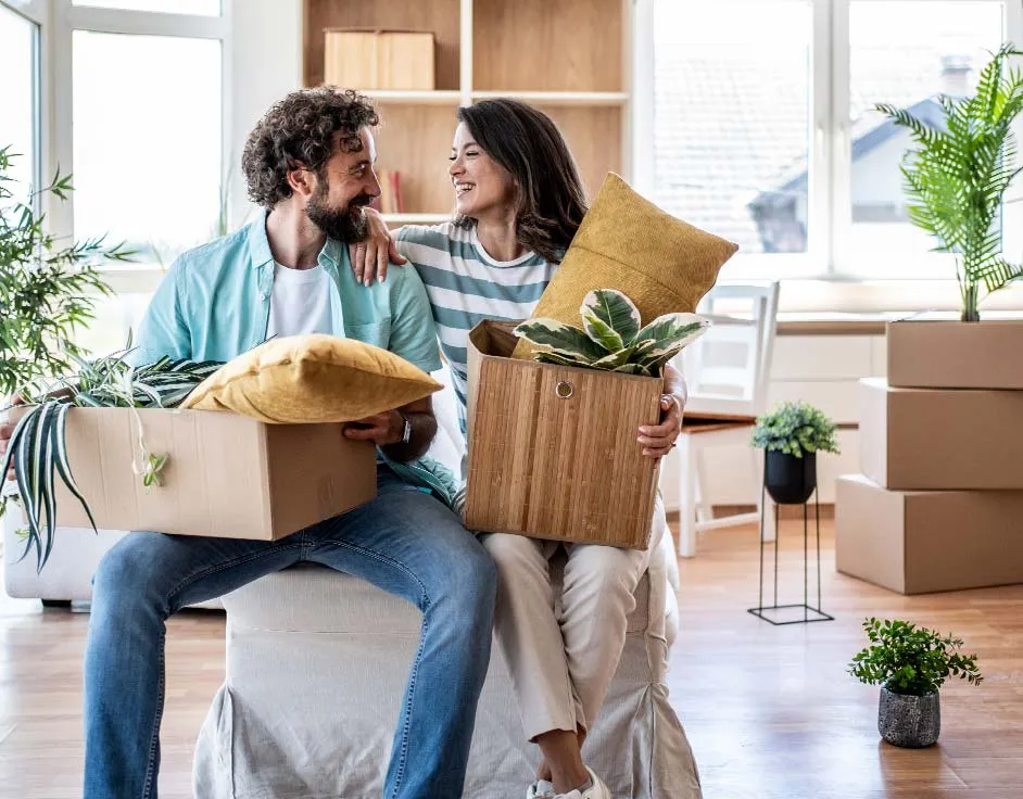 Young adult couple sitting in their new home, holding boxes filled with household items, smiling and looking at each other, starting their life together